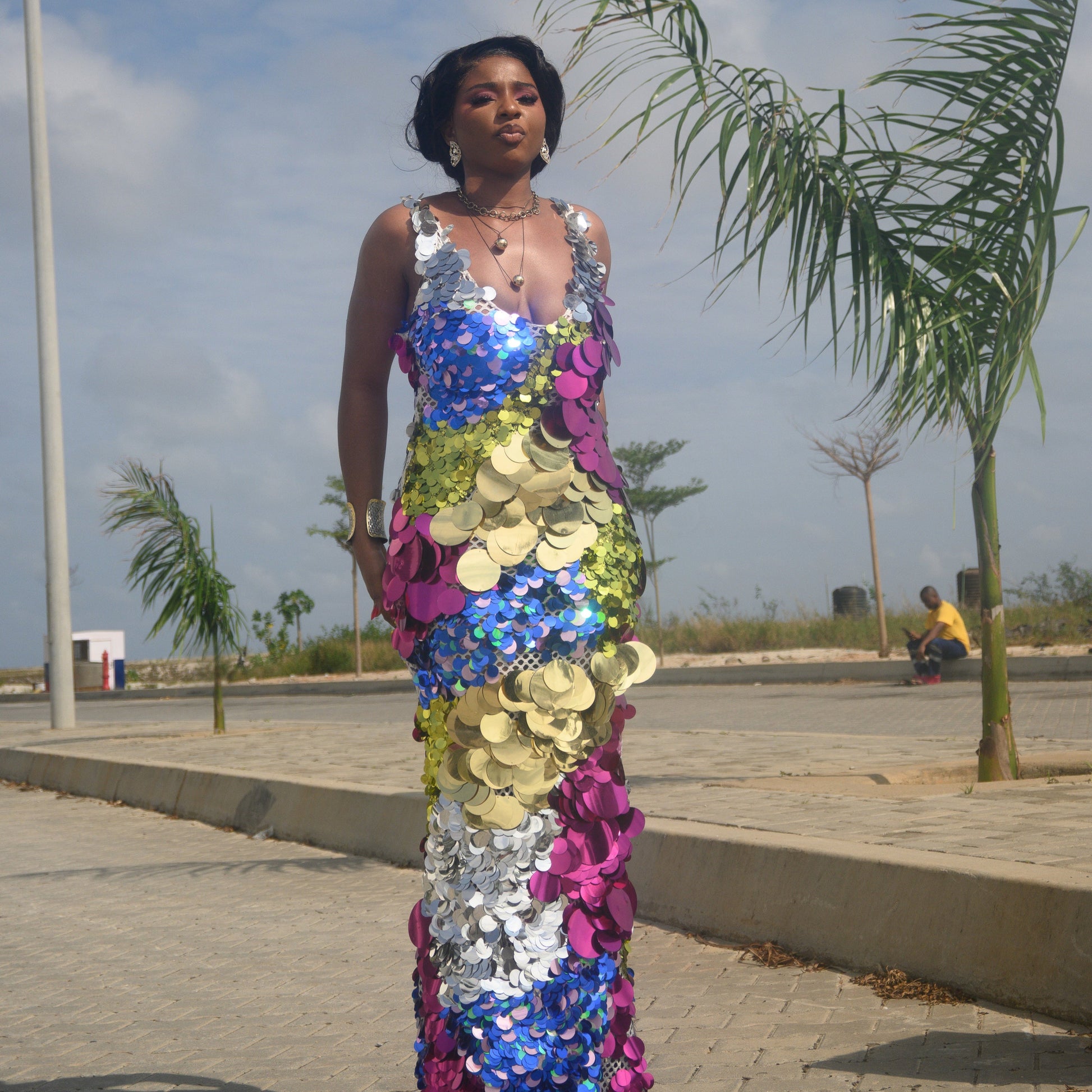 Woman wearing a colorful floral dress standing on a sidewalk with palm trees and a clear sky in the background.